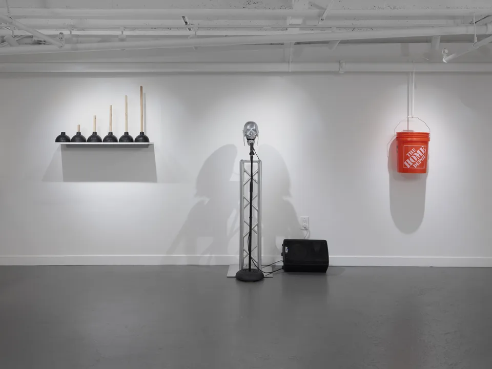 A row of plungers, a dental phantom on a truss, and a hanging orange bucket occupy a white gallery wall.