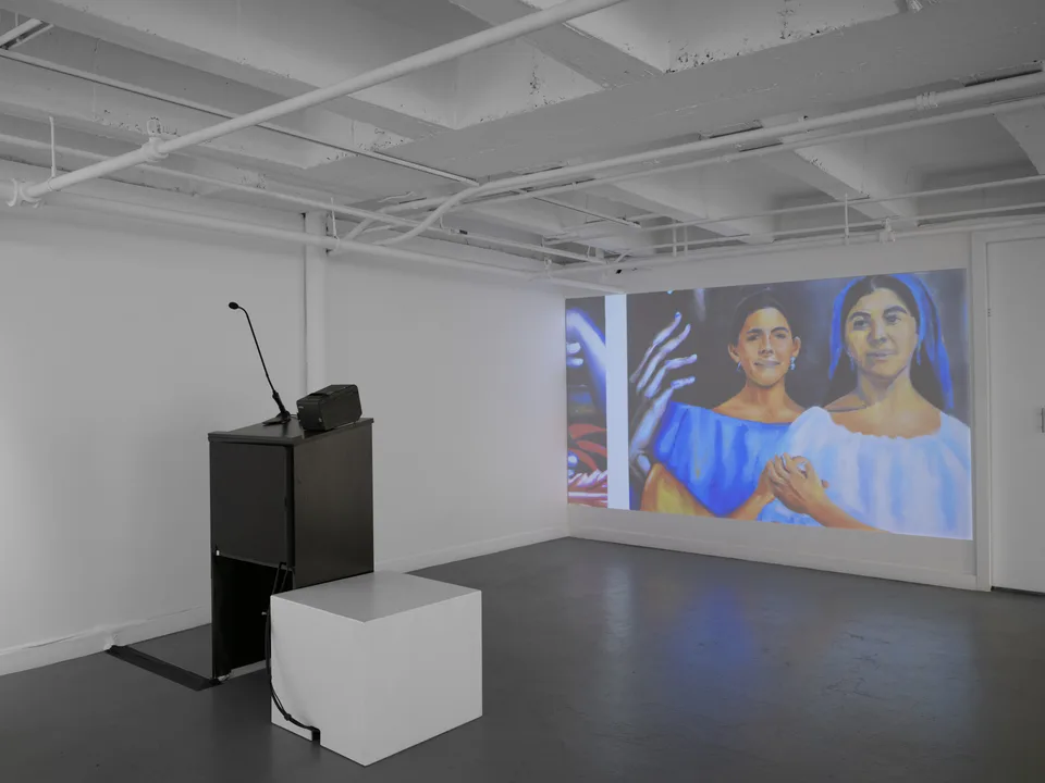 A microphone on a black podium faces a large projected image of two figures in a white gallery.
