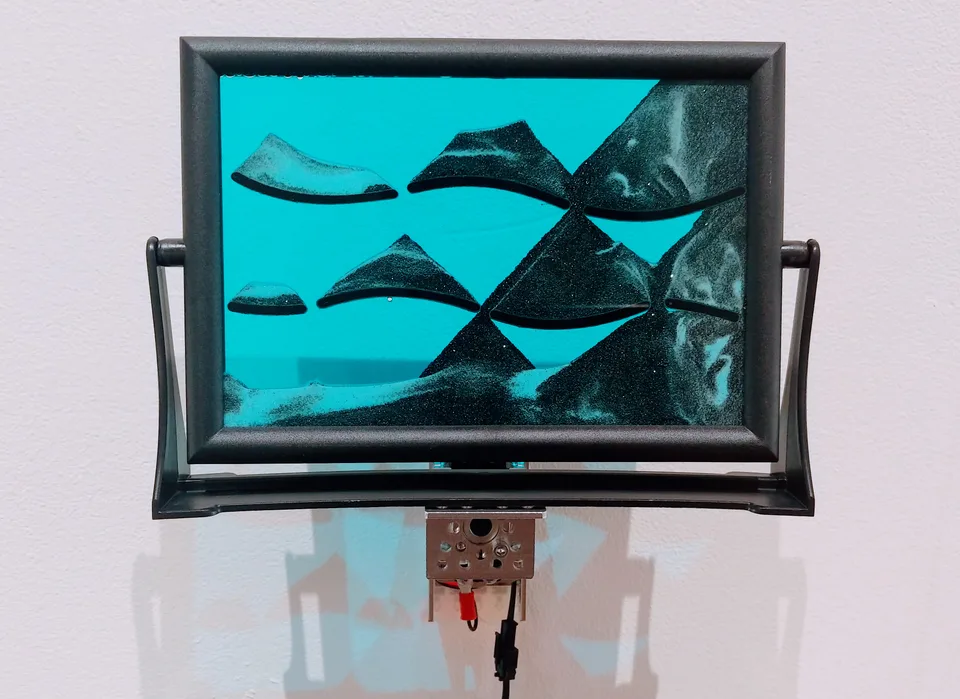 A black rectangular frame holds a blue sand picture with dark sloping forms above a small motorized mechanism.