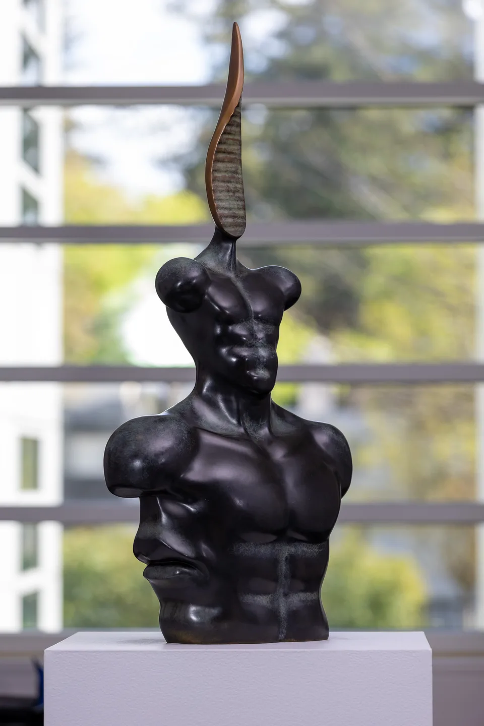 Three-quarter view of the dark bronze bust with a tall blade-like projection beside a window.