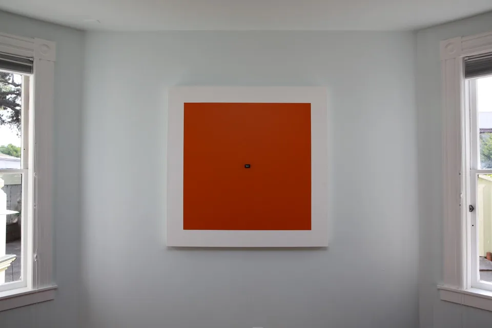 An orange square painting with a small embedded display hanging between two windows