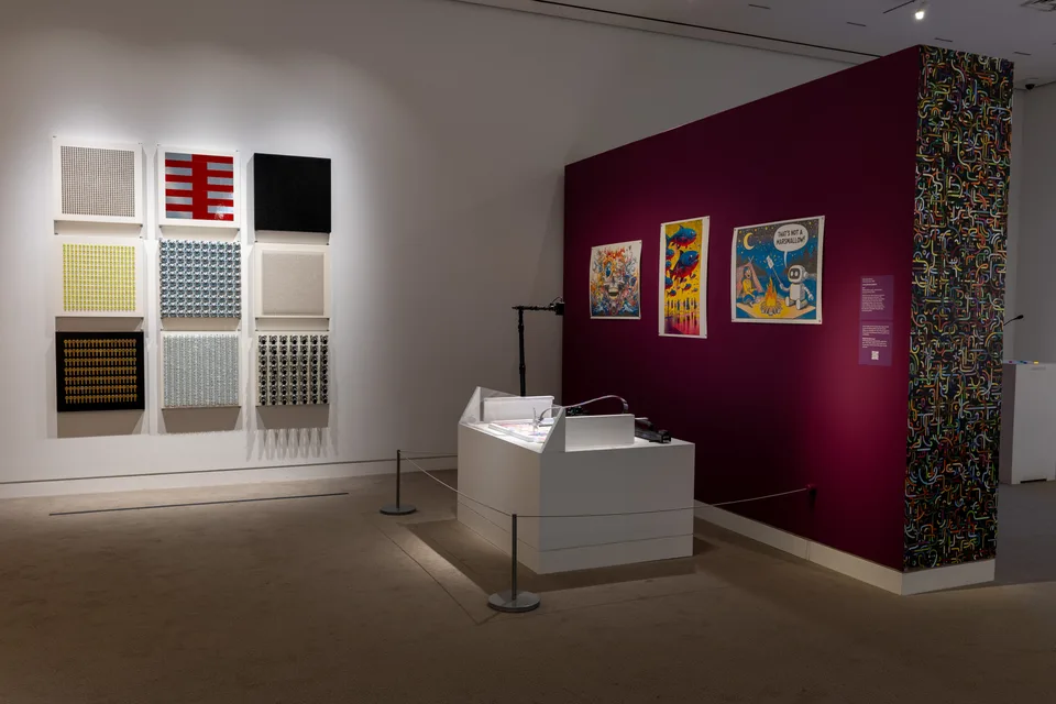 The plotting robot sits in front of a burgundy display wall beside nine panels of repeated found objects.