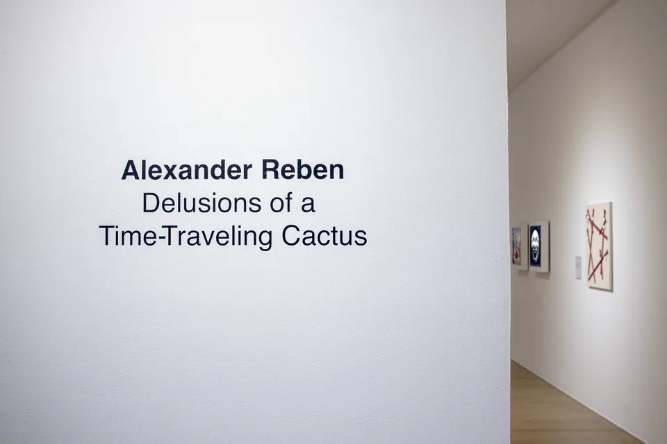 The exhibition title is printed on a white entrance wall, with framed works visible around the corner.