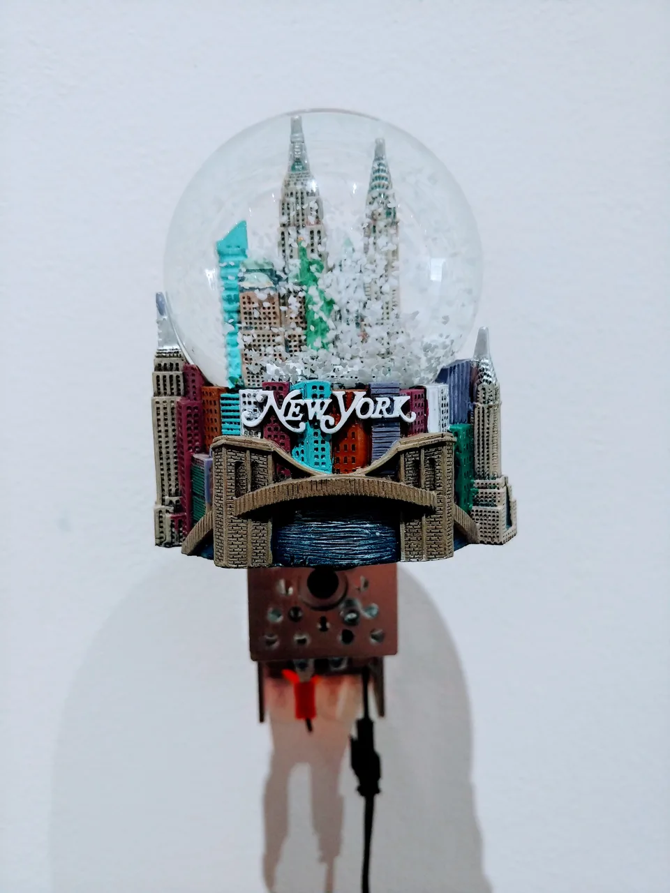 A New York souvenir snow globe with miniature skyscrapers and a sculpted cityscape base is mounted above a motor.
