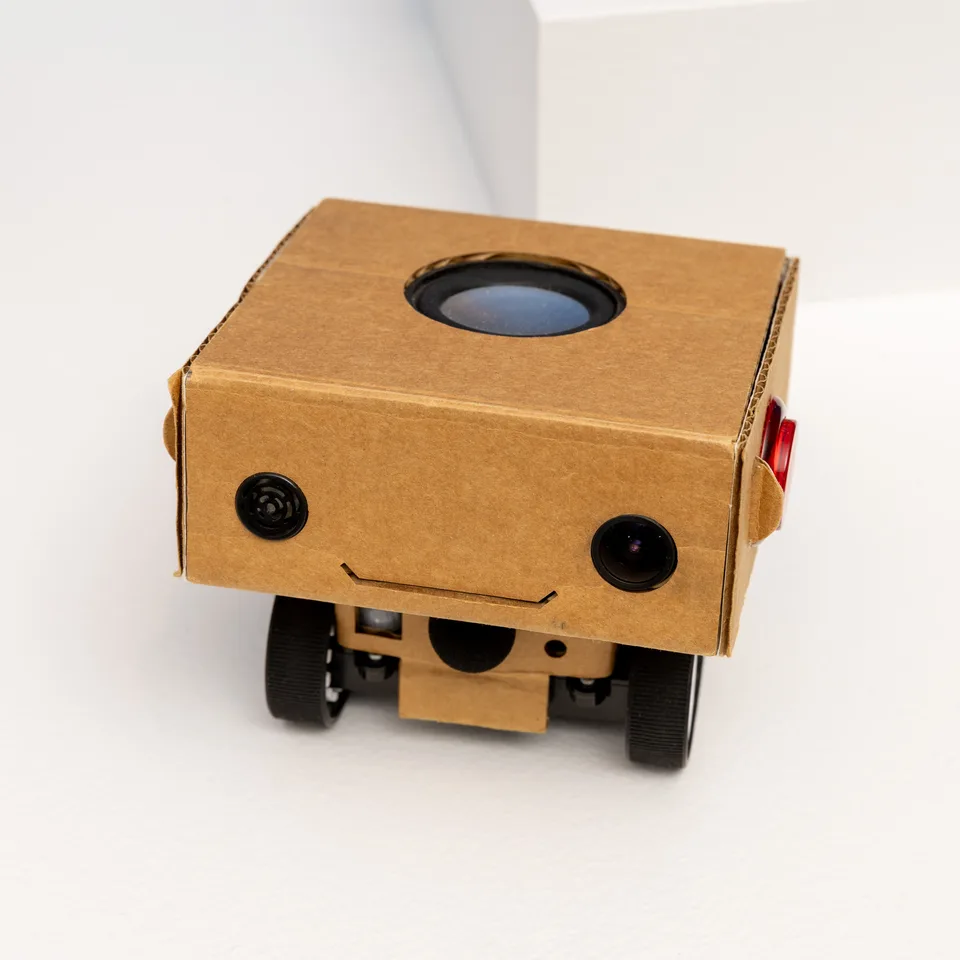 Close-up of a small wheeled cardboard robot with a smiling face and large circular opening on top.