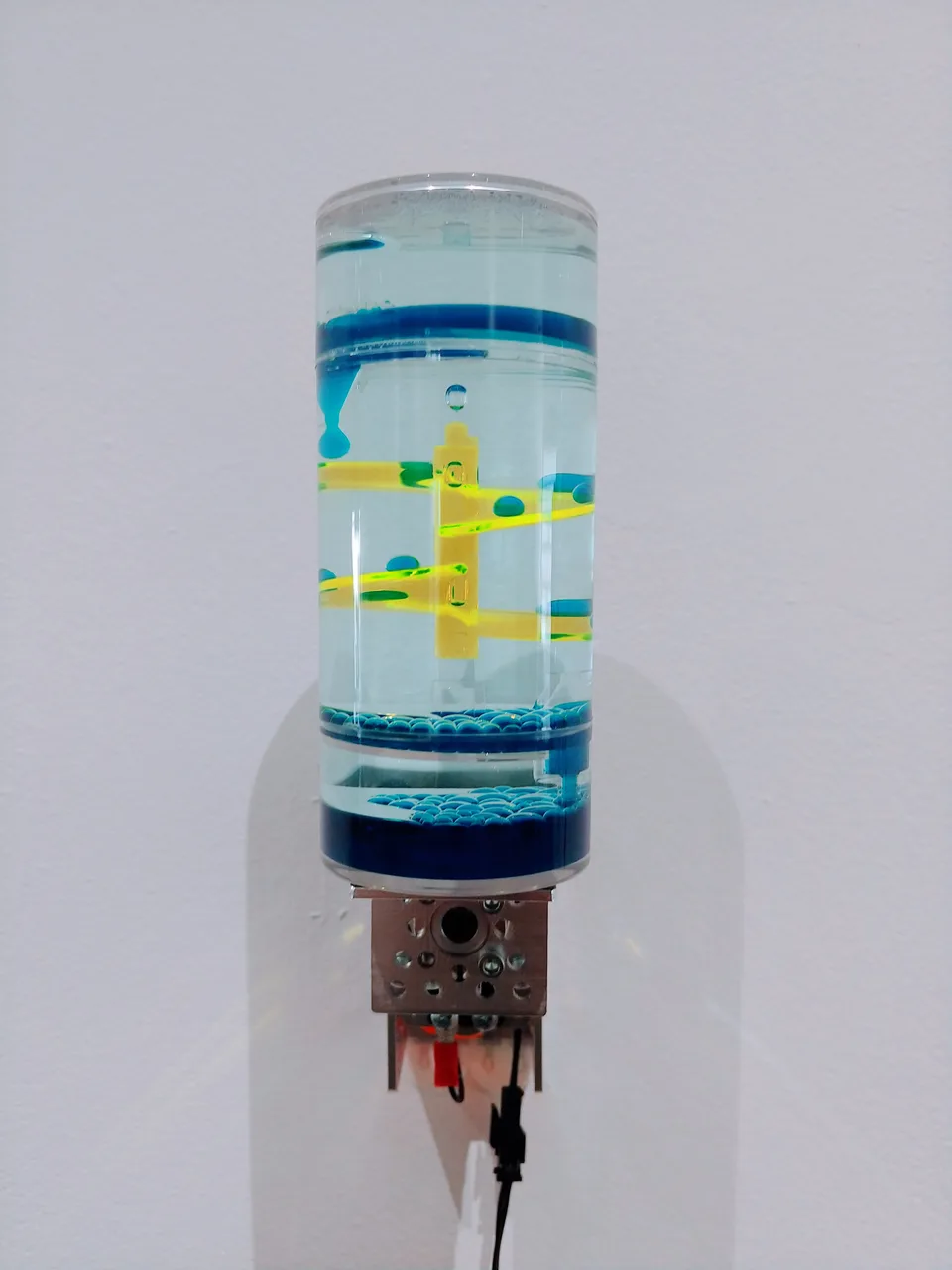 Blue droplets descend across yellow ramps inside a transparent cylindrical toy mounted above a motor.