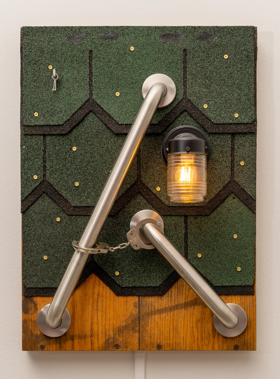 A wall-mounted assemblage combines green roof shingles, two metal grab bars, handcuffs and a glowing light.