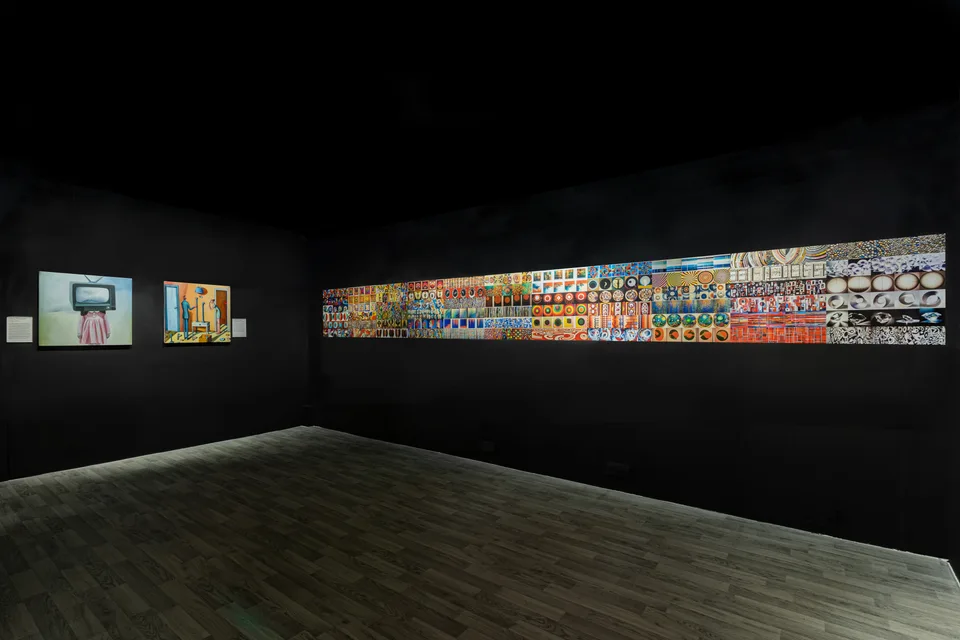 Two square paintings hang beside a long band of small images in a dark gallery with a wooden floor.