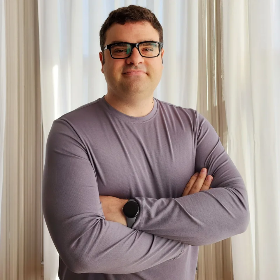 Alexander Reben wearing glasses and a gray shirt, standing with folded arms beside a window