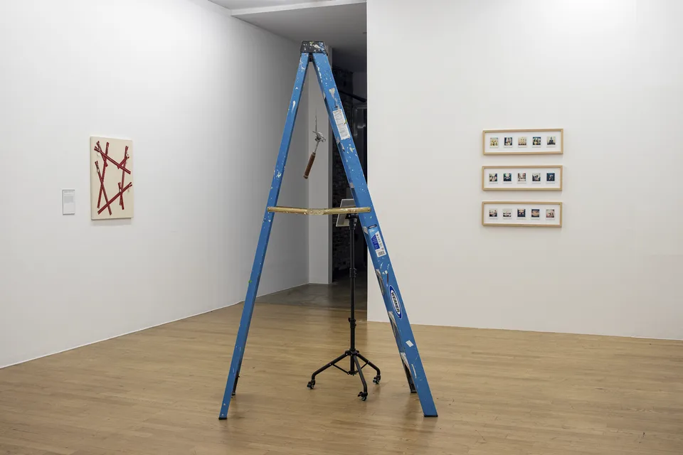 An open blue ladder stands near a canvas of red zippers and three framed rows of Polaroids.