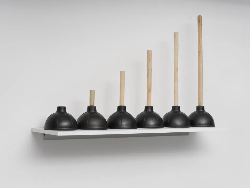 Six black plungers with progressively taller wooden handles stand on a white wall shelf.
