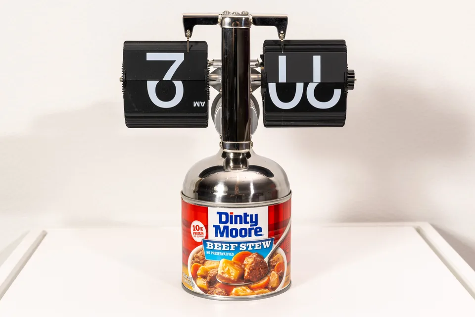 A black split-flap clock mounted on a can of Dinty Moore beef stew.
