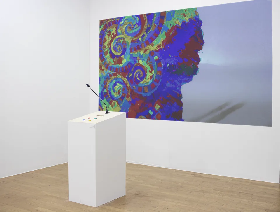 A white pedestal with a microphone and colored controls faces a large projection of a multicolored face.