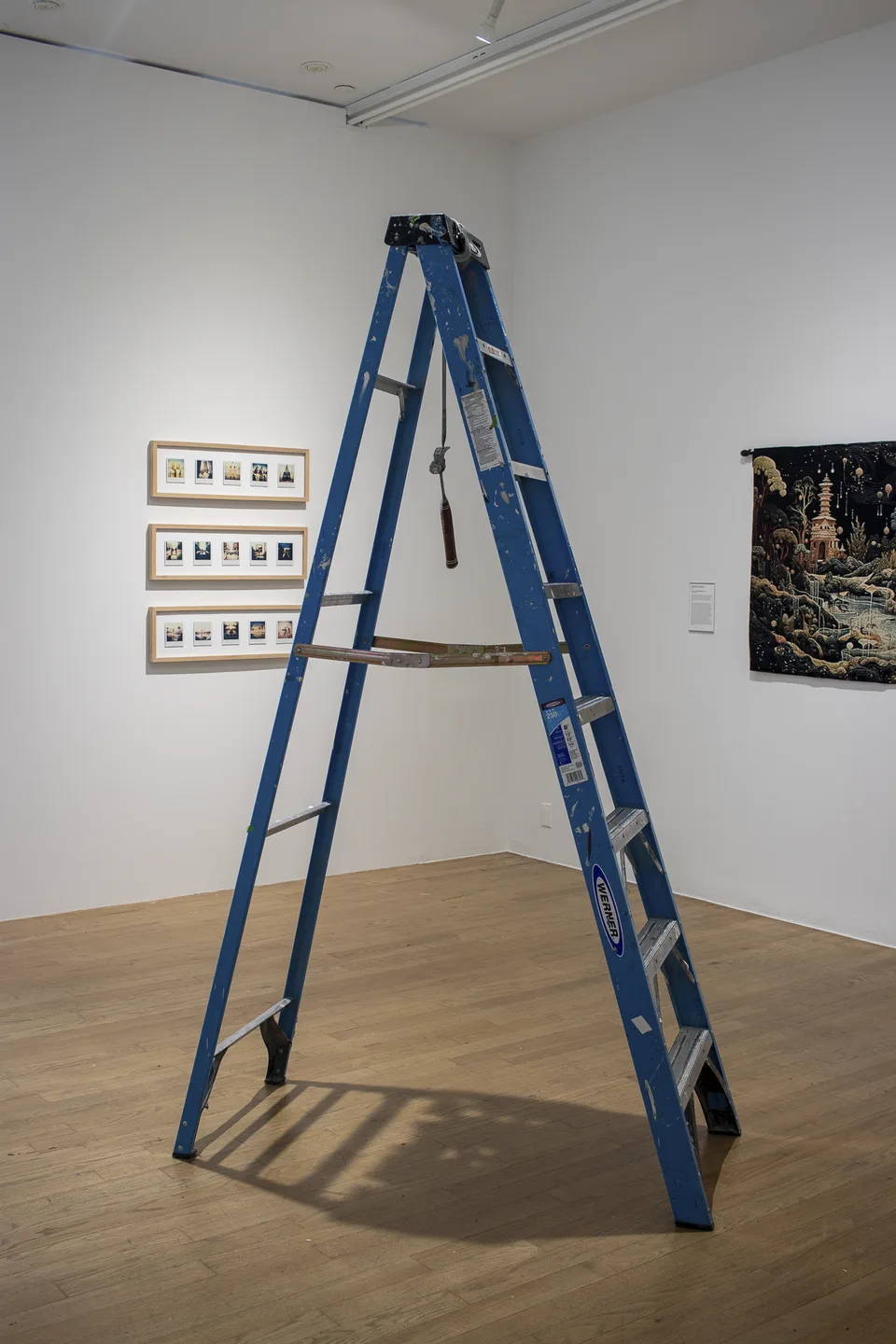 A paint-marked blue stepladder stands open with a hammer suspended from a strap between its sides.