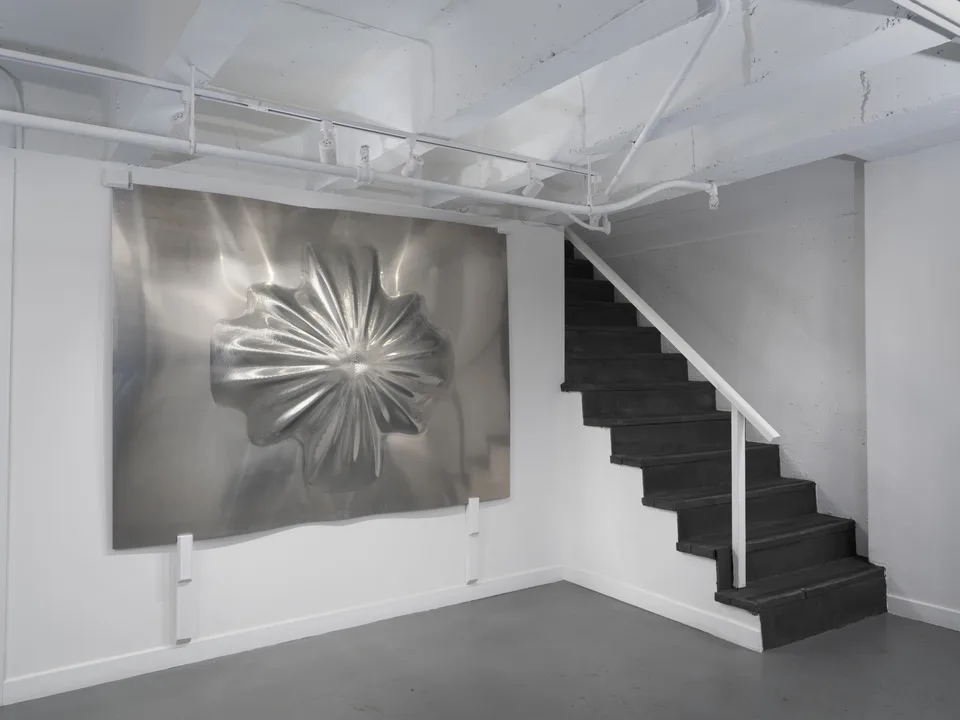 The radial aluminum relief hangs beside a black staircase beneath white ceiling pipes.