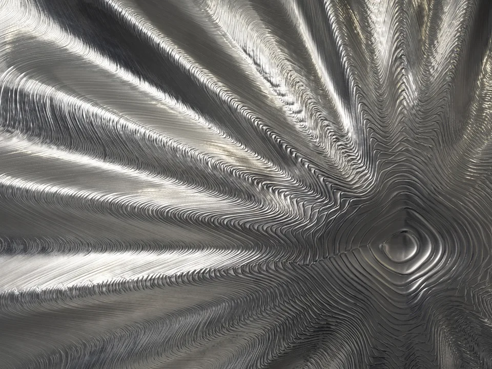 Closely spaced ridges ripple across a reflective aluminum relief toward an off-center circular depression.