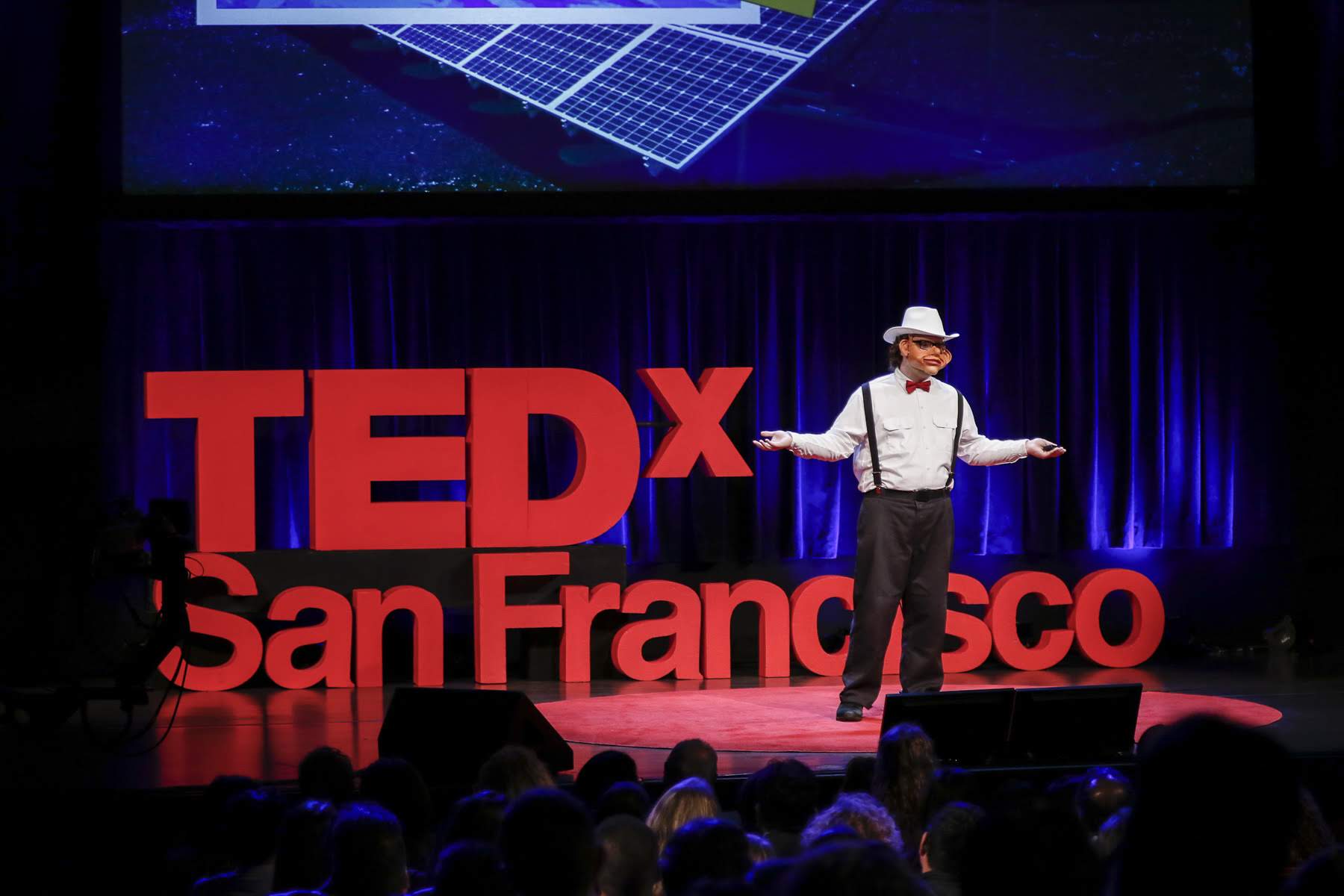 Alexander Reben on stage delivering the AI-written TEDx talk while wearing a robotic face mask